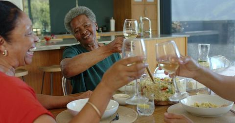 Senior african american friends clinking wine glasses, sharing pasta salad at kitchen table