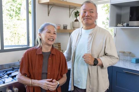 Senior asian couple laughing in sunlit modern kitchen with blue cabinetry