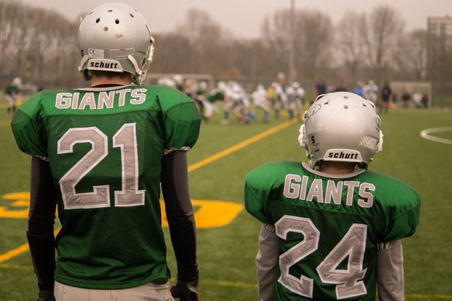 Two young football players wearing green jerseys with their backs to the camera watch a game. The jerseys display the team name 'Giants' and numbers 21 and 24. Both wear helmets, standing on a grassy field with goal posts visible in the distance. Ideal for sports articles, team spirit promotions, and youth athletics content.