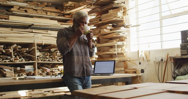 Carpenter taking a coffee break in a woodworking workshop surrounded by wooden planks and tools. Man sipping coffee while standing next to a laptop on a table. Perfect for depicting craftsmanship, small business atmosphere, a balance between manual labor and digital tools.