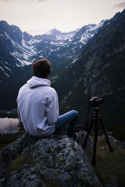 Young photographer is sitting on a rock at sunset, looking at snow-capped mountains. There is a tripod beside him, suggesting he is there for photography. Ideal for themes of travel, adventure, nature photography, and serene landscapes. Suitable for blogs about hiking, photography, or mountain expeditions.
