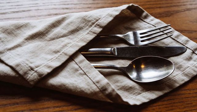 Beige linen napkin pocket holding stainless steel fork knife spoon on rustic wood