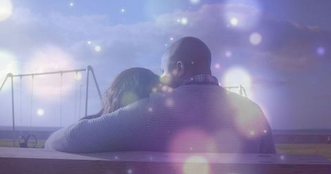 Couple embracing on bench at dusk on waterfront promenade with bokeh lights