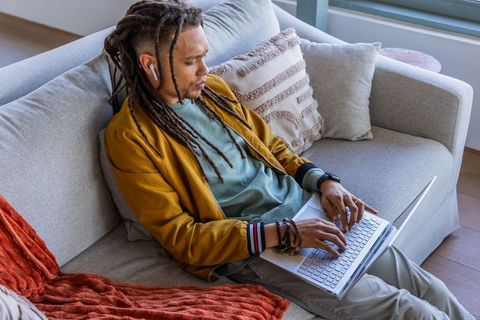 African american man working on laptop on sofa wearing mustard jacket and white earbuds