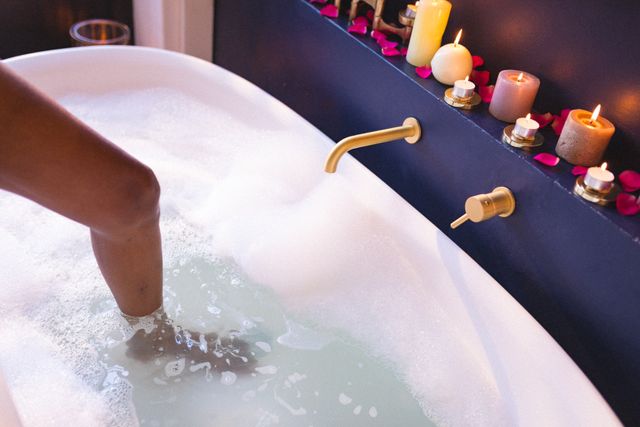 Low section of an African American woman with one leg in a bathtub filled with bubbles. The scene is set with lit candles and rose petals, creating a relaxing and tranquil atmosphere. Ideal for use in articles or advertisements about self-care routines, home spa experiences, relaxation techniques, and wellness practices.