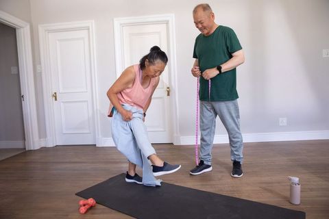 Asian senior couple exercising at home measuring progress with tape, adjusting clothes