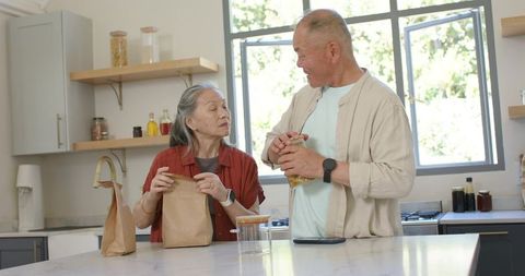 Senior asian couple packing lunch and inspecting pantry jars in bright modern kitchen