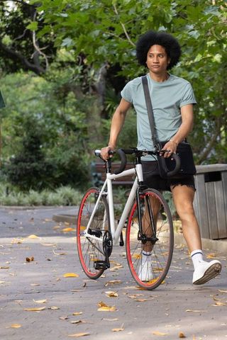 African american man walking white bicycle red rims in urban park carrying messenger bag