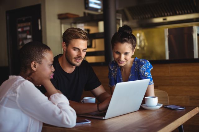 Group of friends using laptop while having coffee in a restaurant. They are collaborating and discussing something on the screen. This image can be used for themes related to teamwork, casual meetings, technology, and social interactions in a relaxed environment.