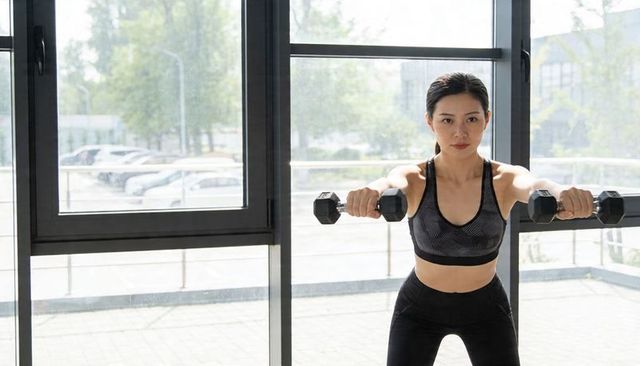 Fit asian woman lifting hex dumbbells in bright gym studio, strength training routine