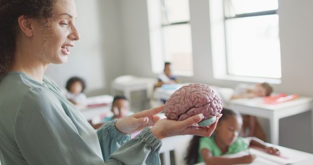 Image of happy caucasian female teacher holding brain model in class of diverse pupils. school education and teaching profession.
