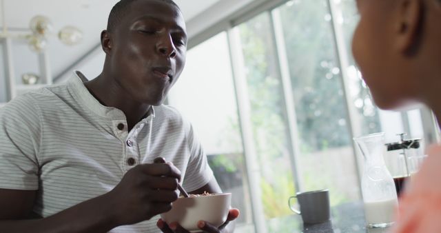 Father sharing breakfast time with daughter at home table, father enjoying a spoonful of cereal. Great for use in ads or articles about family bonding, morning routines, healthy eating, and quality time with children.