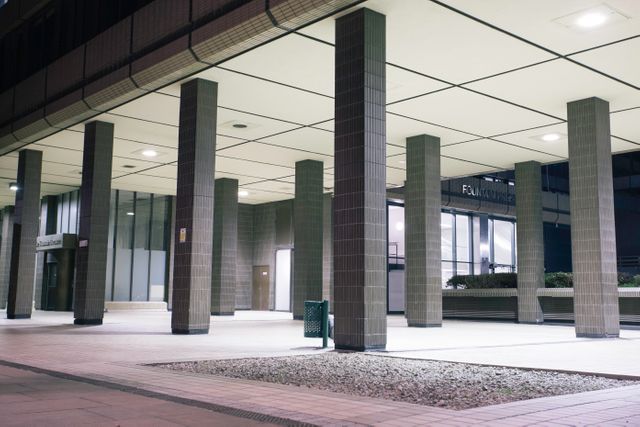 Image showcases a modern urban building entrance at night with rows of pillars providing structural and aesthetic appeal. The strong lines and geometric patterns create a sense of modernism and sophistication. Ideal for use in projects related to urban architecture, city planning, design inspiration, and real estate presentations.