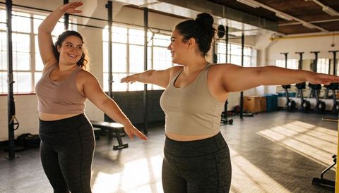 Two women stretching and holding balance in sunlit industrial gym smiling at each other
