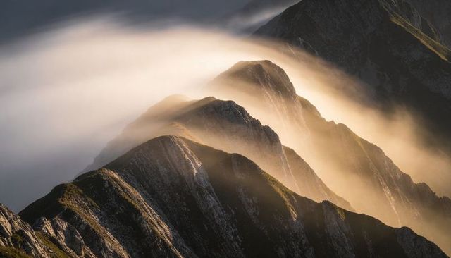 Golden mist cascading over serrated alpine ridge during sunrise, dramatic mountain peaks
