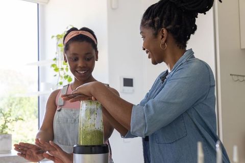 African american mother and teen daughter blending green smoothie in bright home kitchen