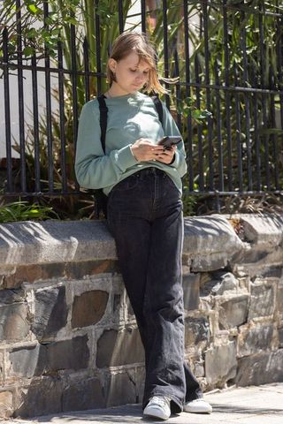 Teenager leaning on wall using smartphone wearing mint sweatshirt and backpack on city sidewalk