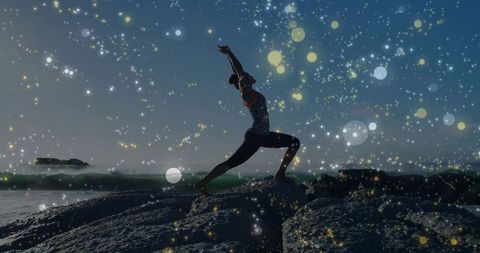Practicing warrior ii pose on rocky coast at sunrise with sparkling bokeh lights
