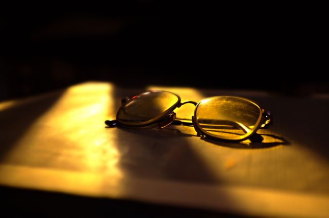 Close-up of a pair of sunglasses resting on a surface with dramatic, golden sunlight creating striking shadows and reflections. Ideal for concepts related to fashion, summer, lifestyle, and vintage aesthetics. Can be used in website banners, social media posts, blog articles, or advertisements.
