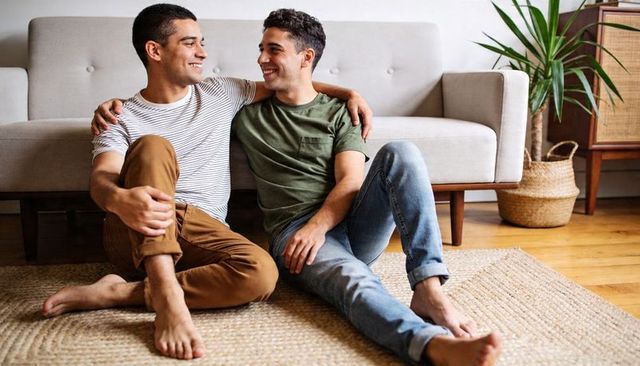 Hispanic male couple sitting on living room floor smiling and embracing on woven rug