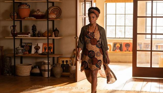 Walking into sunlit artisan studio wearing patterned dress and headwrap, passing pottery
