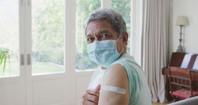 Senior man showing his arm with a medical bandage after vaccination while wearing a face mask at home. Useful for illustrating health campaigns, vaccination drives, and promoting elderly healthcare. Can be used in articles, websites, and brochures about medical health, COVID-19 prevention, and senior wellness.