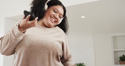 Joyful woman dancing with headphones and remote in bright minimalist living room