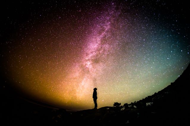 Silhouette of individual standing on a dark hilltop and gazing at vibrant Milky Way galaxy. Colorful night sky filled with stars. Perfect for astronomy enthusiasts, educational materials, websites on space exploration, or inspirational content.