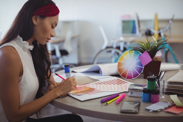 Female entrepreneur working at a desk in a modern office, focusing on design and color palettes. Ideal for use in articles about entrepreneurship, creative workspaces, modern office environments, and professional women in business.