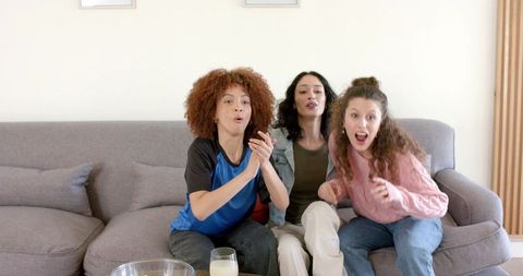 Diverse friends cheering and clapping on gray sofa during cozy game night at home