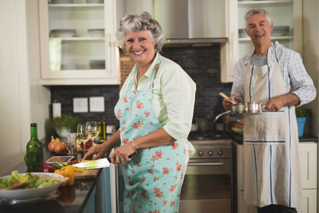 Senior couple enjoying cooking together in a modern kitchen. The woman is chopping vegetables while the man is stirring a pot. Both are wearing aprons and smiling, creating a warm and joyful atmosphere. Ideal for use in advertisements, articles, or blogs about healthy living, retirement, family bonding, and home cooking.