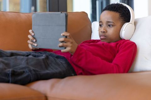 African american boy reclining on couch wearing headphones using tablet at home