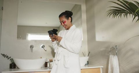 Asian woman enjoying skincare moment with pink eye patches and smartphone in bright bathroom
