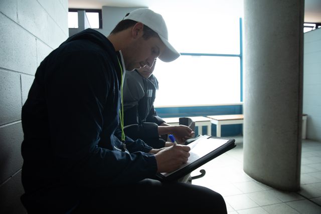 Side view of a male coach and male field hockey players, preparing before a game, sitting in the changing room, holding clipboard talking. Sport game competition.
