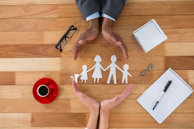 Hands forming a protective gesture around a paper cutout family on a wooden table. Includes items like a coffee cup, glasses, pen, notebook, and key. Ideal for concepts related to family protection, insurance, security, safety, care, support, unity, and teamwork. Useful for advertisements, brochures, websites, and articles focusing on family welfare and protection services.