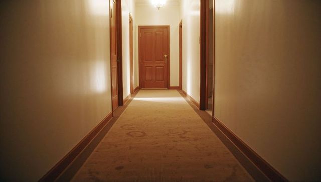 Carpeted hotel corridor leading to closed wooden door with brass handle, warm lighting