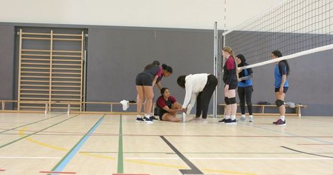 Diverse female volleyball players helping injured teammate on indoor court