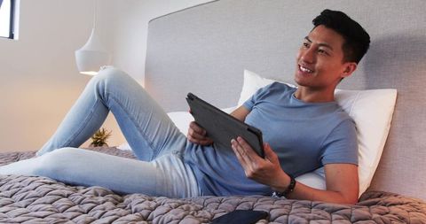 Smiling young asian man relaxing on bed with tablet at home