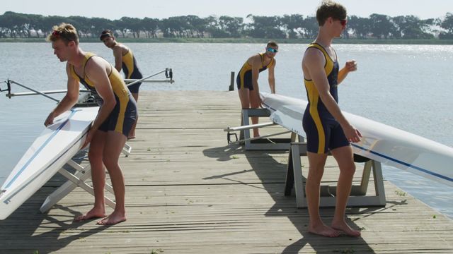 Four young male rowers in uniform prepare their rowing boats on a wooden dock overlooking a calm water body. Each rower participates in boat assembly, demonstrating discipline and teamwork commonly found in the sport of rowing. The sunny day enhances the scene, offering ideal conditions for rowing practice. This image can be used to promote sporting events, highlight teamwork, or feature water-based training practices.