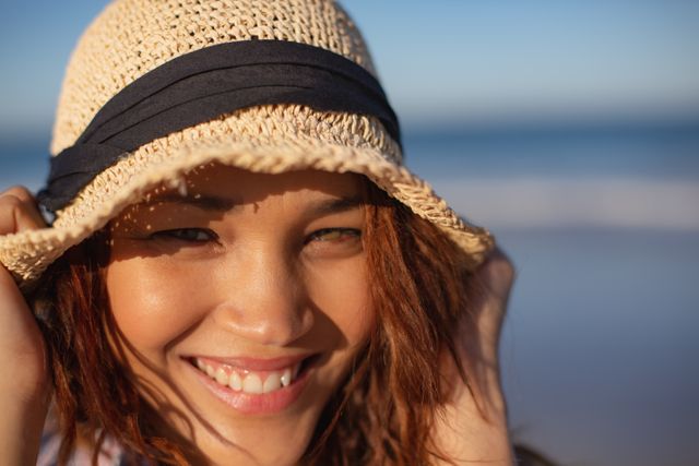 This image captures a close-up of a smiling biracial woman wearing a hat on a sunny beach. Ideal for use in travel brochures, lifestyle blogs, summer vacation promotions, and advertisements focusing on happiness and relaxation.