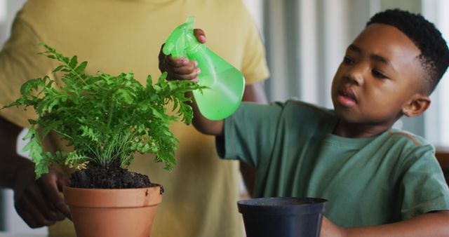 Young boy carefully watering potted plant using green spray bottle, with an adult assisting in background. Useful for topics related to child development, learning through activities, indoor gardening, and connecting with nature. Great for educational materials, family bonding imagery, and sustainable living promotions.