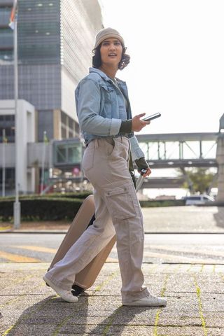 Urban traveler walking with rolling suitcase and smartphone in denim jacket, beanie and pants