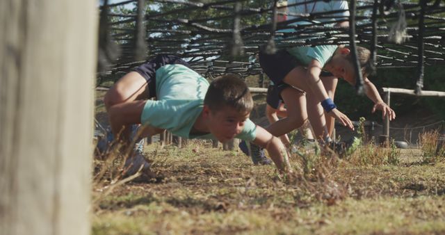 Kids crawling under a net in an outdoor obstacle course, showing teamwork and physical activity. Useful for themes related to children's fitness, outdoor play, teamwork exercises, and adventure activities.