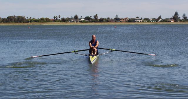 Senior man enjoying tranquil rowing experience on lake in single sculling boat. Ideal for content on fitness, healthy lifestyle, water sports, outdoor recreation, and peaceful nature settings.