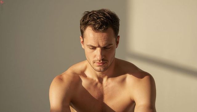 Shirtless athletic man standing in warm studio light looking down contemplative closeup
