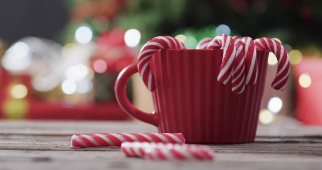 A red mug filled with candy canes is on a wooden table while colorful festive decorations gentle blur in the background. This festive display evokes warmth and holiday cheer, making it an excellent choice for promotion of holiday deals, Christmas advertisements, or social media posts celebrating the holiday spirit.