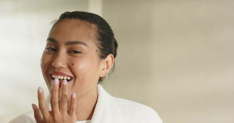 Asian woman smiling while covering mouth with manicured nails wearing white robe
