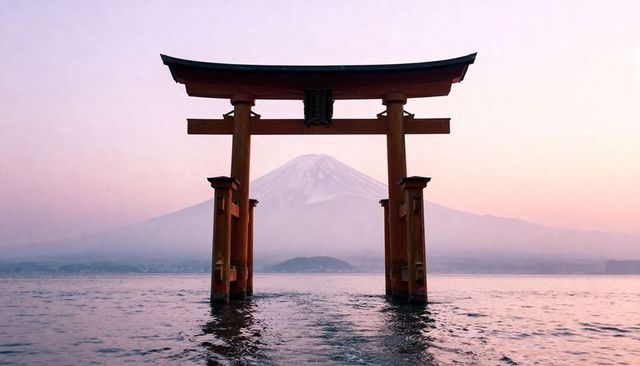 Torii gate framing mount fuji at pastel sunrise over tranquil coastal waters, misty peak