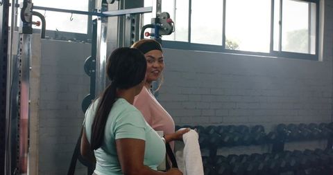 Diverse women chatting after workout in gym with dumbbell rack and cable machine