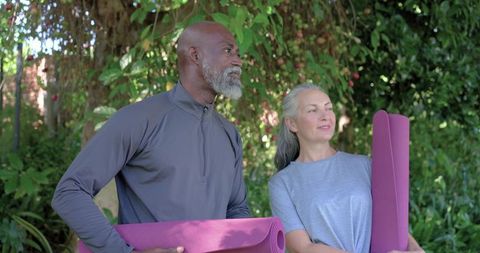 Mature black man and white woman holding magenta yoga mats in sunlit garden practicing yoga